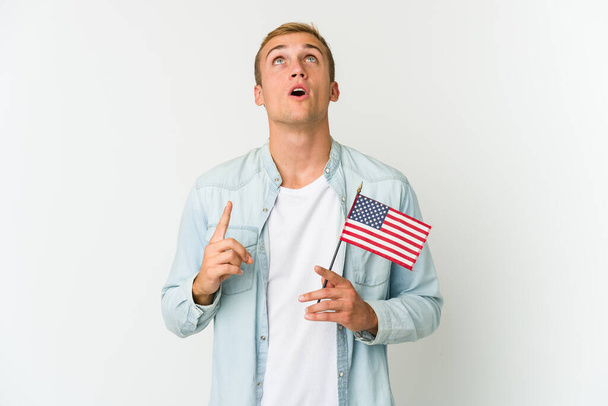 Junger kaukasischer Mann mit einer amerikanischen Flagge auf weißem Hintergrund, die mit geöffnetem Mund auf den Kopf zeigt. - Foto, Bild