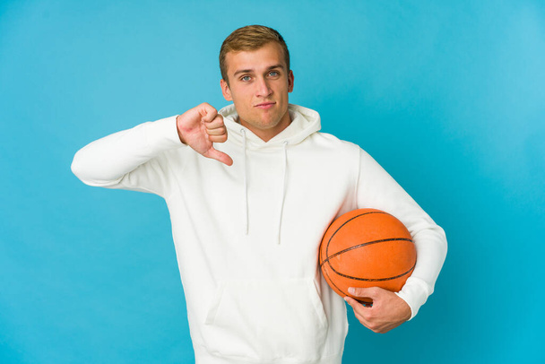 Joven hombre caucásico jugando baloncesto aislado sobre fondo azul mostrando un gesto de aversión, pulgares hacia abajo. Concepto de desacuerdo. - Foto, imagen