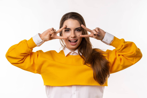Portrait of optimistic brunette girl in casual clothes, showing peace sign over eye and smiling, send positive vibes to you, having faith all be okay, white background. Just be cool about it - Photo, Image
