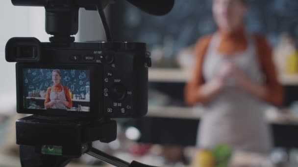 Primer plano enfoque selectivo de la cámara profesional en el trípode recodificación de vídeo de vlogger de comida femenina dando clases de cocina en la cocina - Metraje, vídeo