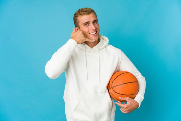 Νεαρός καυκάσιος που παίζει μπάσκετ απομονωμένος σε μπλε φόντο δείχνοντας χειρονομία κινητής τηλεφωνίας με δάχτυλα. - Φωτογραφία, εικόνα