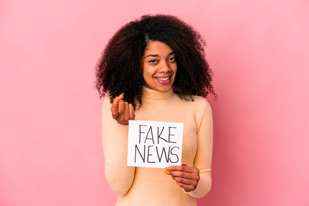 Joven mujer rizada afroamericana sosteniendo una noticia falsa en un cartel señalándote con el dedo como si invitaras a acercarte. - Foto, Imagen