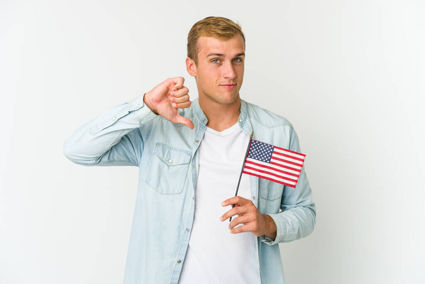 Junger kaukasischer Mann mit einer amerikanischen Flagge auf weißem Hintergrund, die eine ablehnende Geste zeigt, Daumen nach unten. Konzept der Uneinigkeit. - Foto, Bild