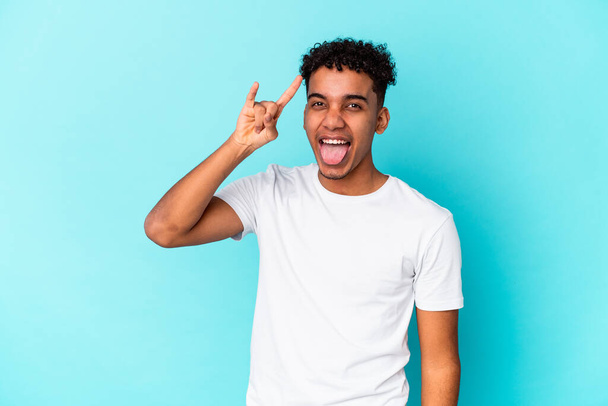 Νεαρός αφροαμερικανός σγουρός άνδρας απομονωμένος σε μπλε που δείχνει ροκ χειρονομία με δάχτυλα - Φωτογραφία, εικόνα
