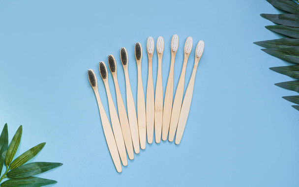 Set of eco friendly wooden bamboo toothbrushes on a blue background with tropical leaves. Oral cavity care. The concept of environmentally friendly disposal of toothbrushes. Flat lay. top view.  - Фото, изображение