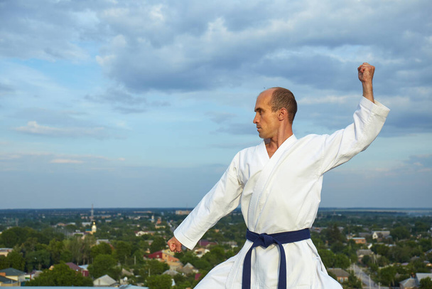 Sportman met blauwe gordel voert formele karate oefeningen uit op stadsachtergrond - Foto, afbeelding