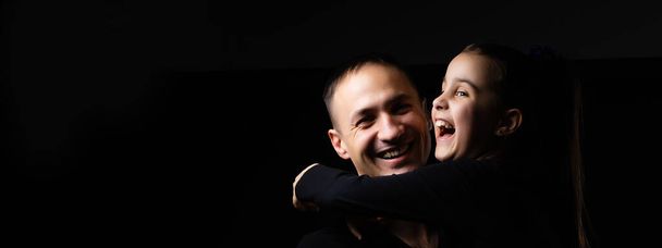 Young dad with a cute emotional daughter on a dark background. Place for your text. - Φωτογραφία, εικόνα