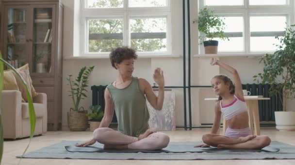 Tracking shot van fit jonge moeder en dochtertje in sportkleding zitten gekruiste benen op yoga mat en stretching samen in de woonkamer - Video