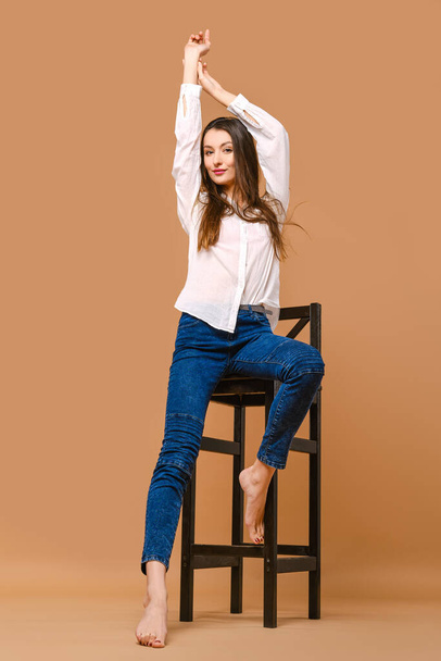 Young woamn with bare feet and in casual outfit posing at tall wooden stool in studio - Фото, зображення