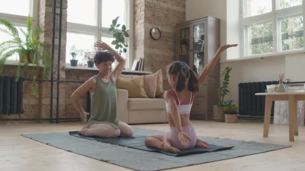 Suivi Slowmo de joyeuse jeune mère et sa jolie petite fille assise dans sa pose en face de l'autre sur un tapis de yoga et étirant leurs bras derrière leur dos - Séquence, vidéo