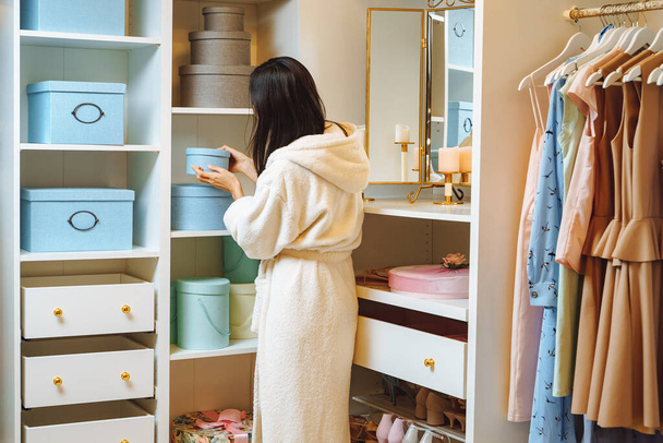 Young woman in a bathrobe taking a blue box from a wardrobe at home. Fashion and storage concept. - Foto, Imagen