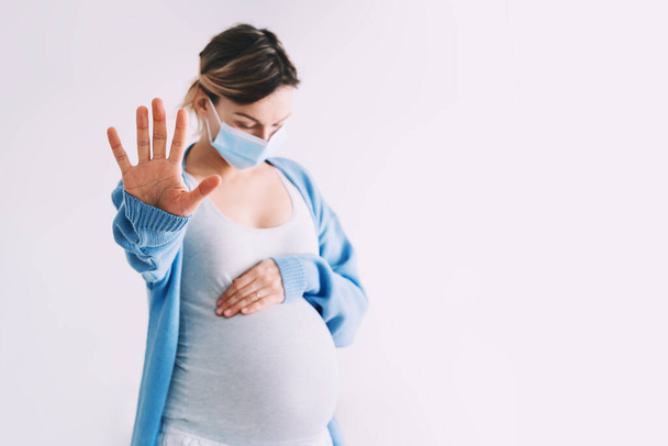 Pregnancy and childbirth during Infections. Pregnant woman in medical mask hugging belly showing STOP gesture. Protection and prevention during viruses, mother care of her and unborn baby health. - Photo, image