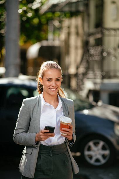 Ευτυχισμένη νεαρή επιχειρηματίας χρησιμοποιώντας το τηλέφωνο στο δρόμο κατά τη διάρκεια του φρένου του καφέ - Φωτογραφία, εικόνα