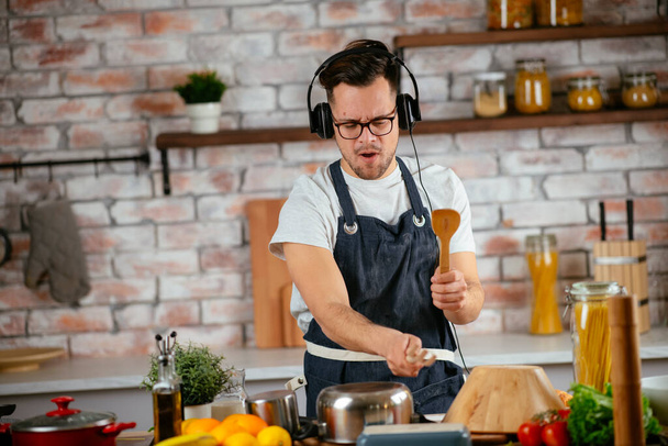 Ευτυχισμένος άντρας με ακουστικά που ακούει μουσική και χορεύει ενώ μαγειρεύει στην κουζίνα. - Φωτογραφία, εικόνα