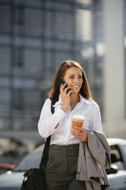 Mulher bonita que vai trabalhar com telefone celular em sua mão ao caminhar perto do prédio de escritórios. - Foto, Imagem