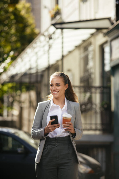Fiatal és gyönyörű üzletasszony használja a telefont kávészünetben, miközben sétál az utcán - Fotó, kép