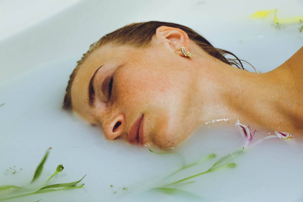 beautiful girl lying in a bath with milk water with flowers and leaves, spa tender young woman posing sensually touching on a light background copy space closeup portrait - Valokuva, kuva