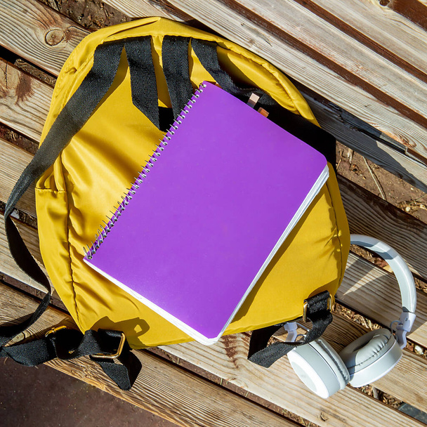 yellow backpack, purple school notebook and white headphones lie on a wooden bench - Zdjęcie, obraz