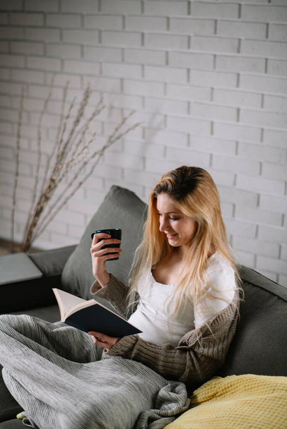 Молода красива дівчина читає книжку на дивані вдома. Жінка читає книгу в затишній квартирі
. - Фото, зображення