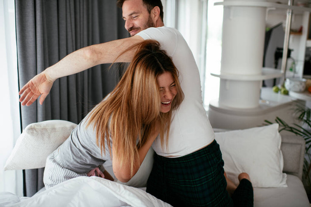 ベッドの上で枕と戦う若いカップル。幸せなカップルが家で楽しんでいる - 写真・画像