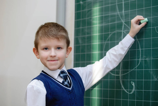 Schuljunge in Uniform schreibt mit Kreide auf eine Schultafel. Grundschulbildung. Selektiver Fokus. - Foto, Bild