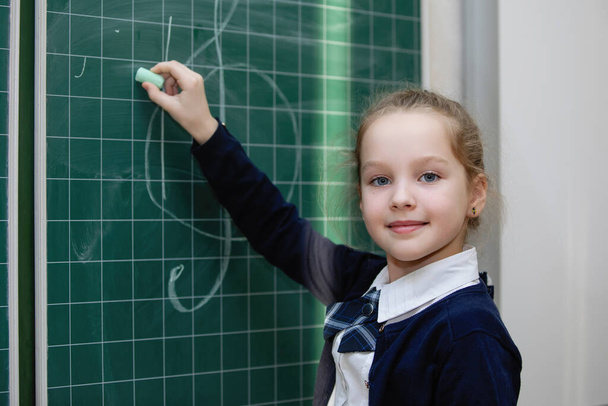 Schulmädchen in Uniform schreibt mit Kreide an eine Tafel. Grundschulbildung. Selektiver Fokus. - Foto, Bild
