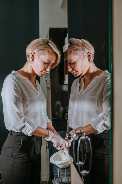 Professional hairdresser young blond woman prepairing hair dye at hair salon standing near mirror - Photo, Image