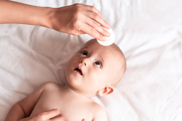 Onderwerp Hygiëne reinigingsdoekje, zuiver, schoon. Moeders hand veegt het gezicht van pasgeborenen af met een wit wattenschijfje. Baby 's gezicht afvegen. Zorg voor een schone en zachte huid. - Foto, afbeelding