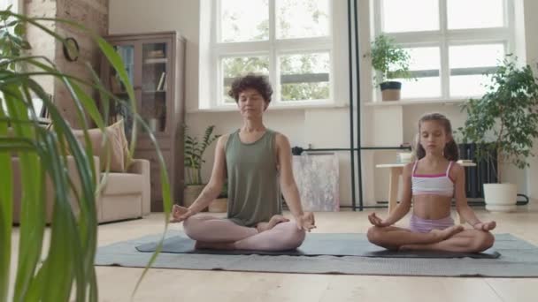 Tracking shot de joven madre e hija pequeña en ropa deportiva sentada sobre pose de loto en esterilla de yoga y meditando en casa - Metraje, vídeo