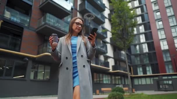 Mulher vestida formalmente andando pela rua em um distrito de negócios com café na mão e usando um smartphone - Filmagem, Vídeo