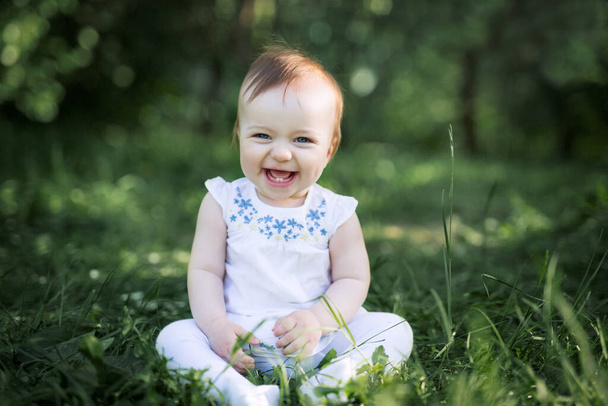 公園の緑の芝生の上に赤ちゃんが腰を下ろして笑う。最初の2本の歯は赤ん坊が微笑むときに見える。健康な赤ん坊は笑う。気をつけて,  - 写真・画像