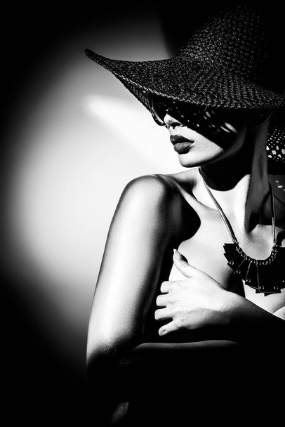 Έννοια μόδας. Πορτρέτο μιας όμορφης γυναίκας με μεγάλο καπέλο και γυαλιά ηλίου σε έντονο φως αντίθεσης. Γυναικείο μοντέλο που φοράει αξεσουάρ ποζάρει στο στούντιο. Επαγγελματικό μακιγιάζ. Μαύρο και άσπρο μονόχρωμο - Φωτογραφία, εικόνα