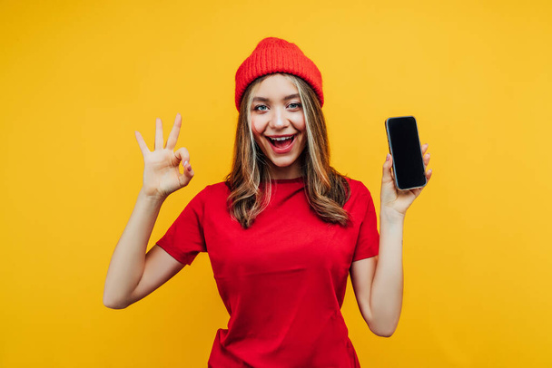 Close up photo of a pretty girl on a yellow background in the studio, she holds a phone with a black screen in her hand and the other hand shows okay. She is dressed in bright red clothes. - Φωτογραφία, εικόνα