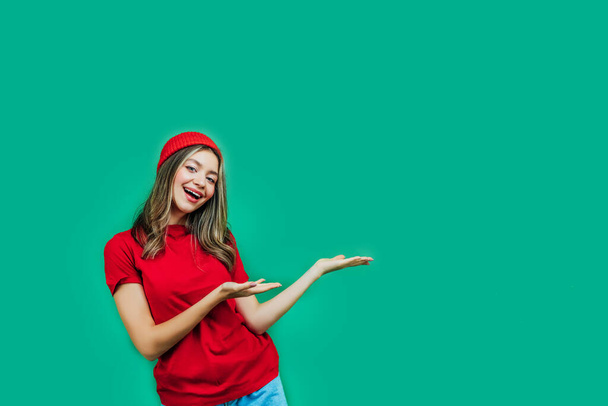 Green background. Pretty girl in red clothes on a green background with space for text, she shows her hands in the direction of free space. Copy the place. Place for text. - Fotografie, Obrázek