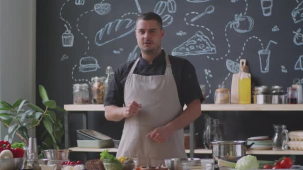 Профессиональный шеф-повар в фартуке, нюхающий хлеб, бросающий авокадо и выступающий перед камерой во время съемок видео для кулинарного блога на кухне - Кадры, видео