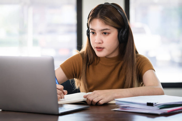 Joven estudiante asiática lleva auriculares inalámbricos concentrados escribir en el cuaderno para estudiar el lenguaje en línea ver y escuchar al conferenciante, webinar a través de video llamada e-learning en casa, educación en línea - Foto, Imagen