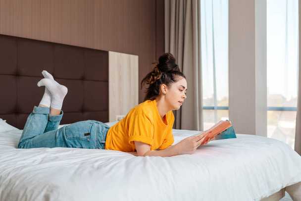 Olvasás és kikapcsolódás. Egy fiatal csinos nő fekszik az ágyon és újságot olvas. Ablakok a háttérben. Beltéri, beltéri. - Fotó, kép