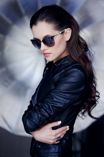 Confident and sexy young brunette woman posing in black leather jacket and sunglasses, on a creave silver background. - Photo, Image
