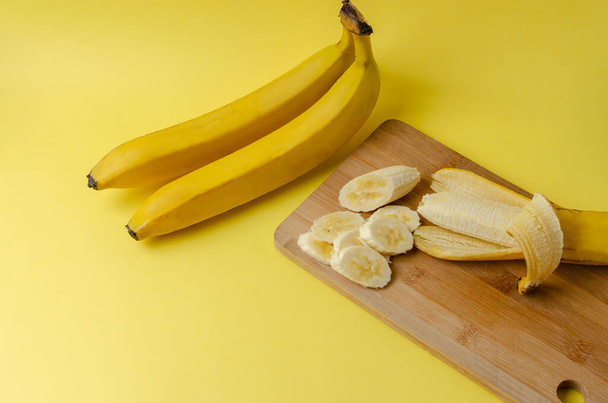 Two bananas and sliced banana on a wooden cutting board on a yellow background. Trending color Pantone of year 2021 Illuminating. Side view minimal still life. - Photo, Image