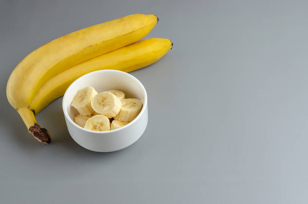 Two bananas and slices in a white plate on a gray background. Trending color Pantone of year 2021 Illuminating and Ultimate gray. Side view minimal still life with copy space. - Foto, imagen