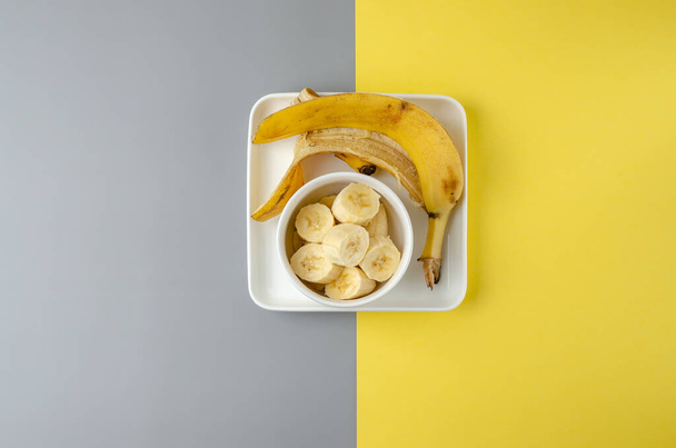 Slices of bananas and banana peel in a white plate on a gray and yellow background. Trending color Pantone of year 2021 Illuminating and Ultimate gray. Top view minimal still life - Fotografie, Obrázek