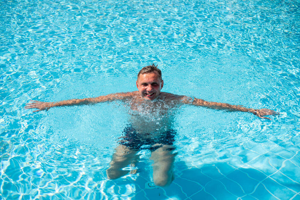 przystojny facet pół nagi wesoły mężczyzna uśmiechający się śmiejąc się w błękitnym basenie wodnym. Zdjęcie przystojnego uśmiechniętego mężczyzny w basenie w letniej scenerii - Zdjęcie, obraz