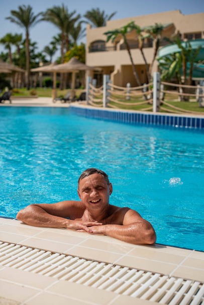 przystojny facet pół nagi wesoły mężczyzna uśmiechający się śmiejąc się w błękitnym basenie wodnym. Zdjęcie przystojnego uśmiechniętego mężczyzny w basenie w letniej scenerii - Zdjęcie, obraz