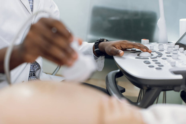 Healthcare, ultrasound concept. Close up cropped shot of hands of male African doctor, screening of pregnant woman by ultrasound scan, pushing buttons on control panel. Focus on hand on buttons - Foto, imagen