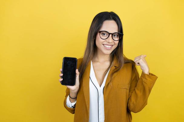 Fiatal gyönyörű nő visel blézer felett elszigetelt sárga háttér mosolyog, rámutatva a fénymásolás tér és a kezében egy telefont a másik kezét - Fotó, kép