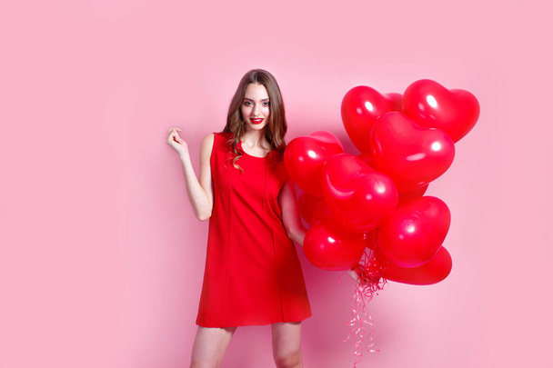 Valentines Day. Beautiful elegant young woman in red dress with heart shape red air balloons on pink background. - Fotografie, Obrázek