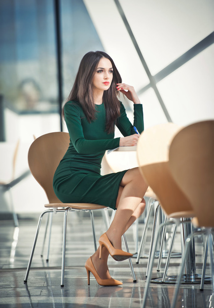 Модная привлекательная девушка в темно-зеленом платье, сидящая на стуле с надписью, снимок в помещении. Современный городской пейзаж. Модное художественное фото чувственной дамы в стеклянной и стальной декорациях. Девушка с высокими каблуками за столом
 - Фото, изображение