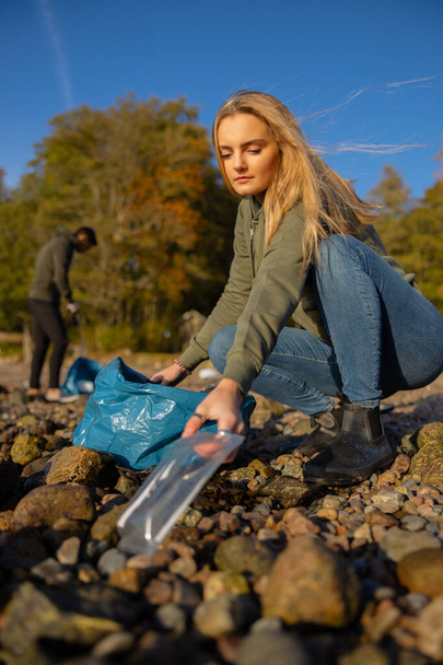 Mujer joven y seria recogiendo plástico en la playa rocosa - Foto, imagen