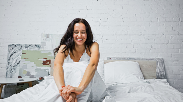 cheerful brunette woman with closed eyes stretching while sitting in bed - Photo, image