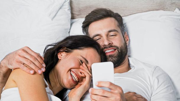 glimlachende man met behulp van mobiele telefoon terwijl knuffelen opgewonden vriendin in bed - Foto, afbeelding